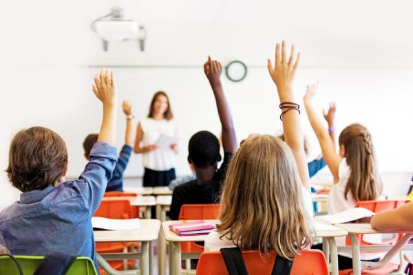 Students raising hands in classroom with teacher standing at front